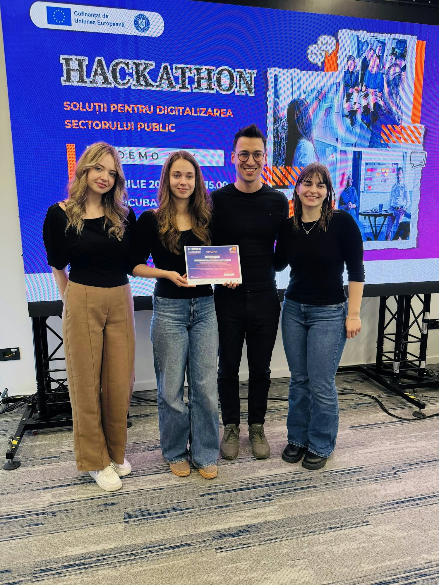 Four young people, three women and one man, stand smiling in front of a large screen at a hackathon event. Two of them hold up a diploma together. The screen behind them reads 'HACKATHON — Soluții pentru digitalizarea sectorului public' (Solutions for digitalising the public sector) and displays an EU co-funding logo alongside event photos