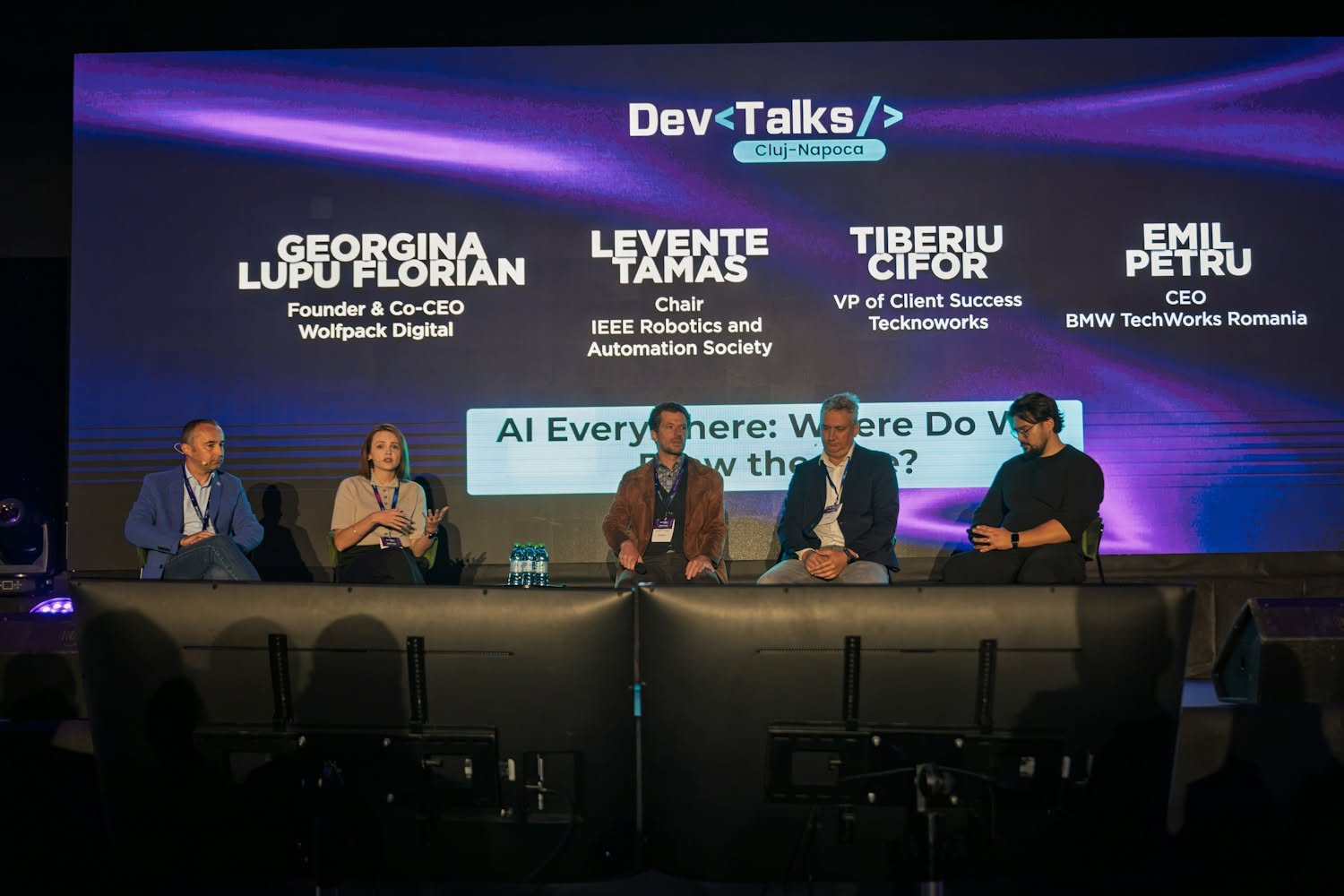 Georgina Lupu Florian, Founder and co-CEO of Wolfpack Digital, speaking on the “AI Everywhere: Where Do We Draw the Line?” panel at DevTalks Cluj-Napoca, seated on stage with four other speakers in front of a large screen showing the panellists’ names.