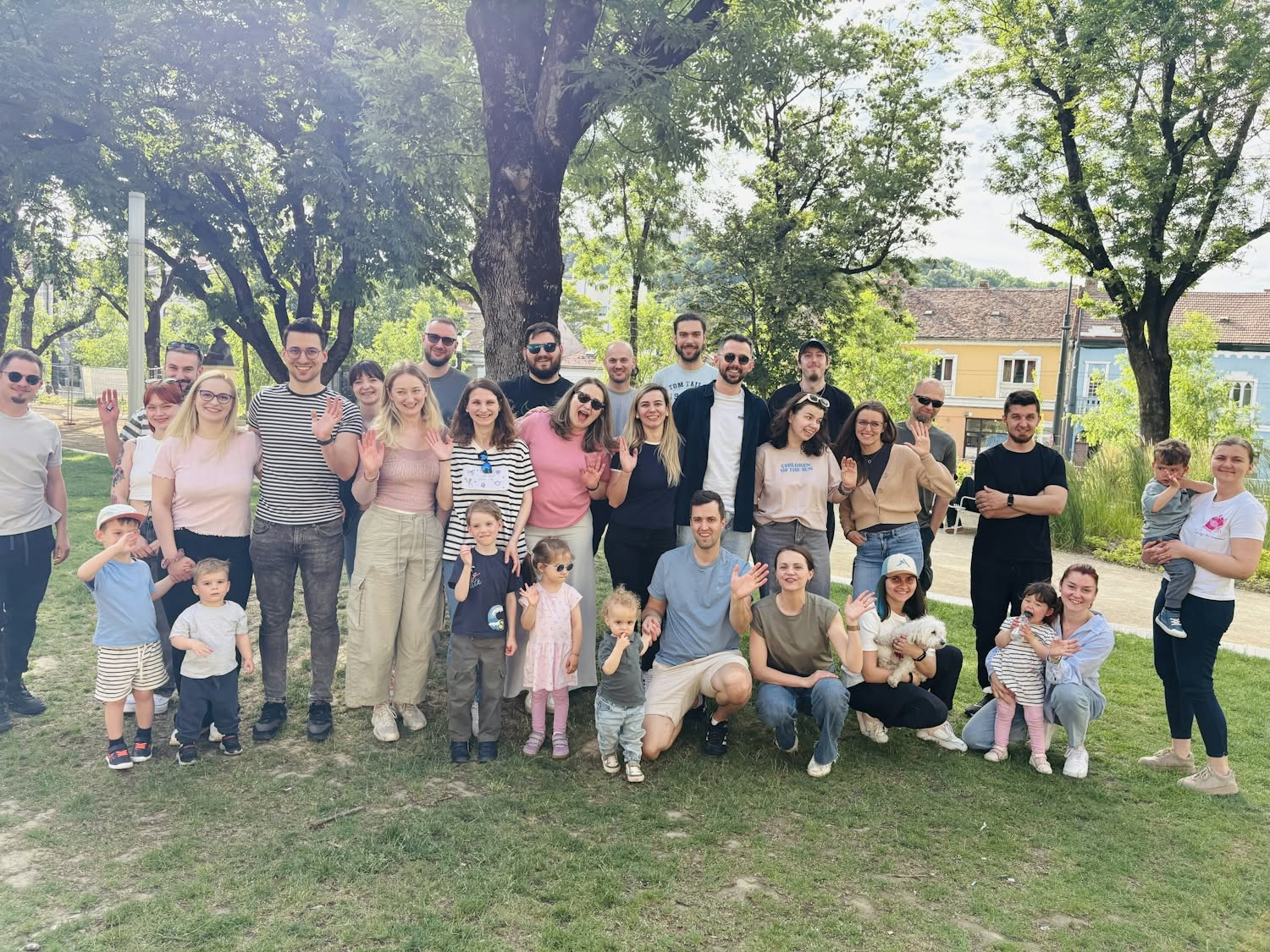 The Wolfpack Digital team and their families gathered outdoors in a Cluj-Napoca park for a summer get-together, smiling and waving at the camera beneath tall trees.