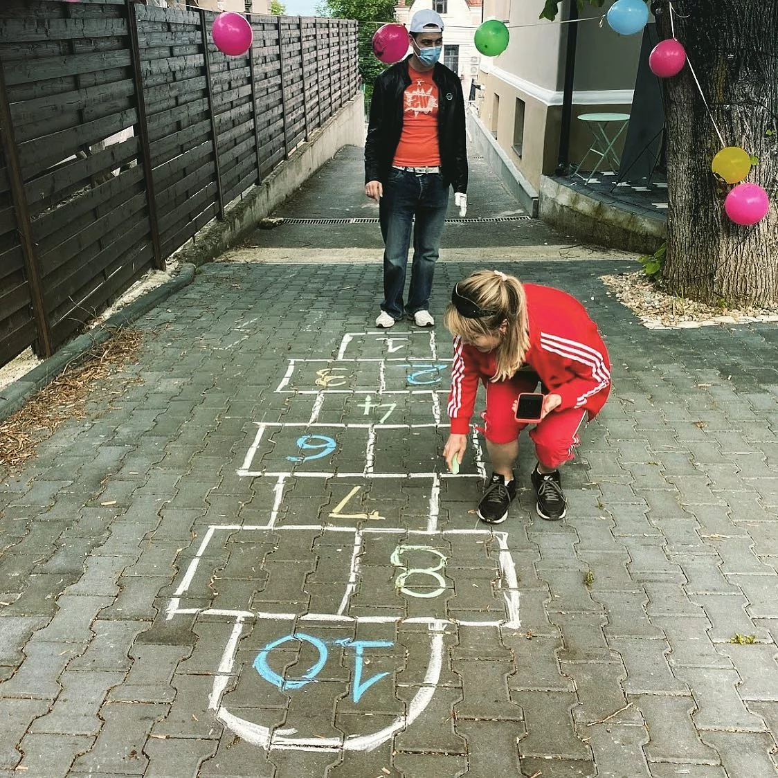 Person in a red tracksuit crouching to play chalk-drawn hopscotch in a paved courtyard, a nostalgic Q2 2021 theme