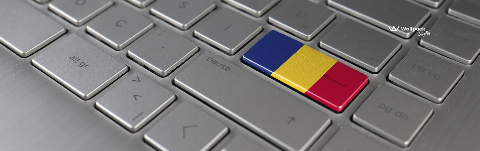 Close-up of a laptop keyboard with one key replaced by the Romanian flag in blue, yellow, and red, with the Wolfpack Digital logo in the top right corner.