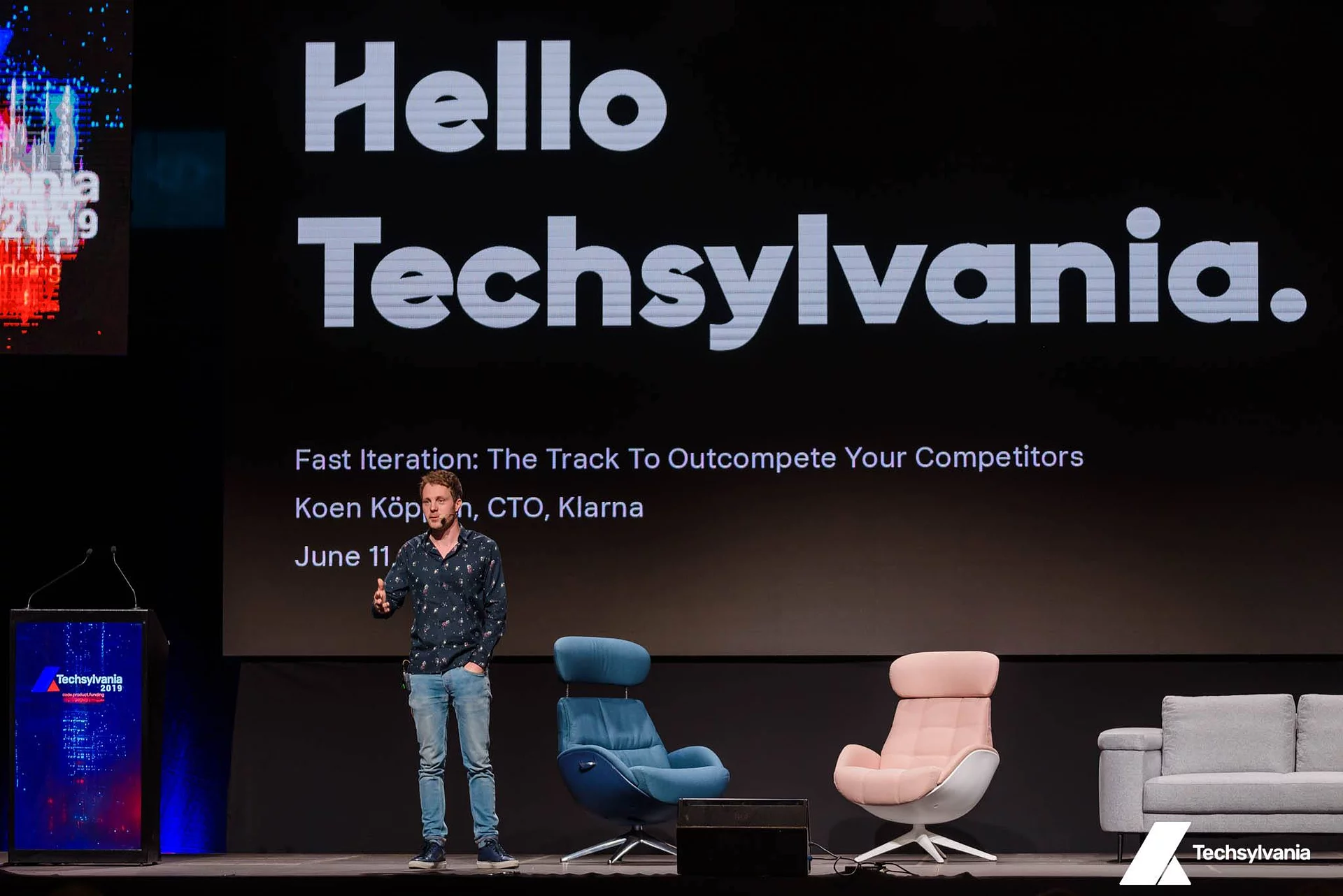 Speaker on stage at Techsylvania in front of a large screen reading 'Hello Techsylvania,' with armchairs and a sofa arranged beside him.