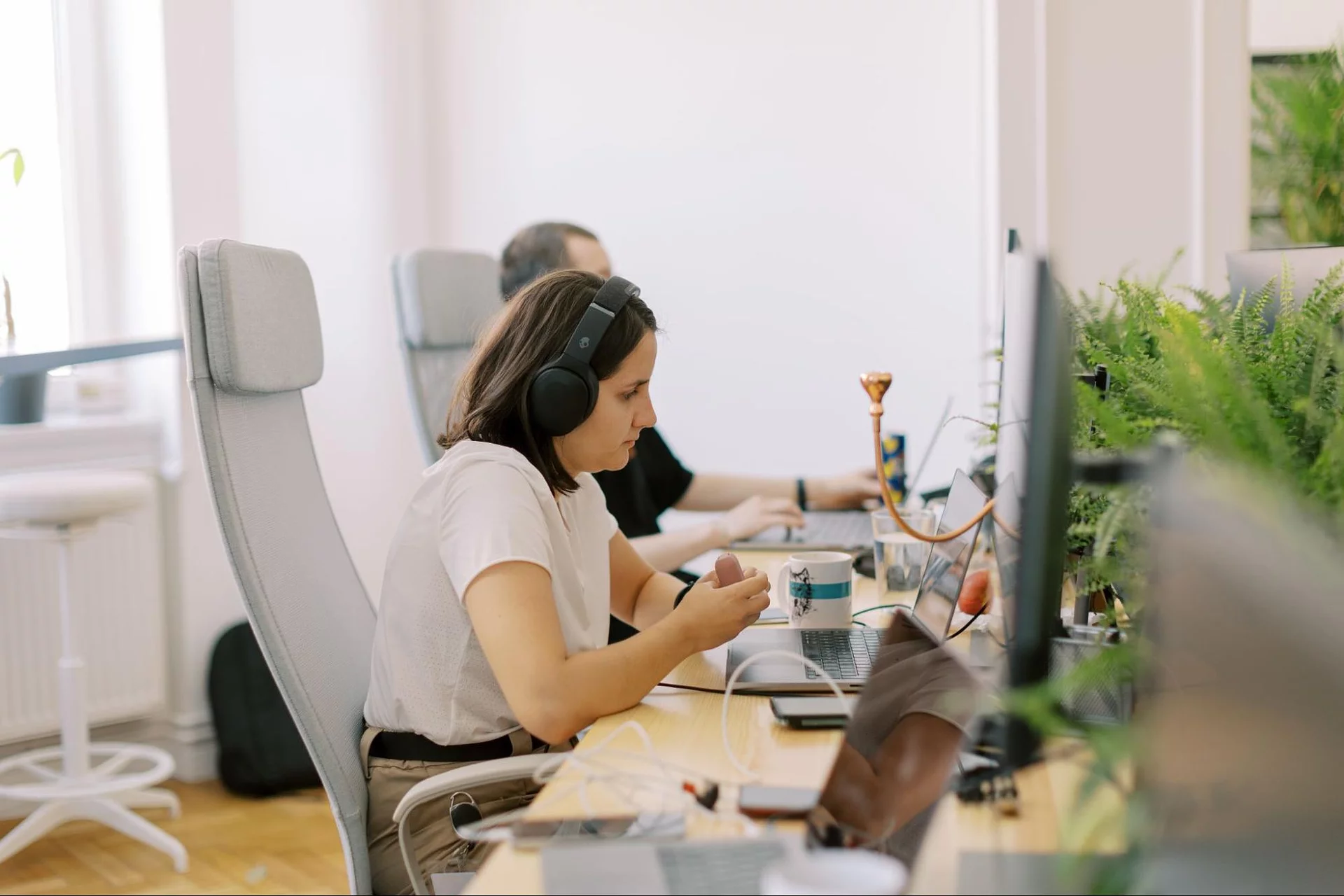 Team members working at desks in a modern office, a woman in the foreground wearing headphones focused on her screen beside green plants